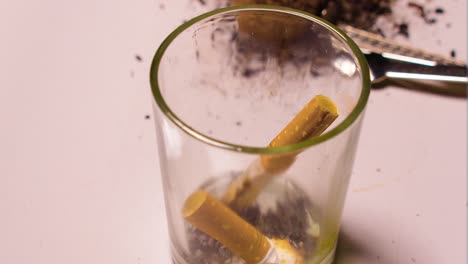 person holding a lit cigarette flicking into a glass using it as an astray as they are smoking inside their home on a white table bad habit two cigarettes tossed away finished close up