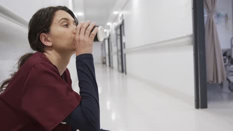 Doctora-Caucásica-En-Cuclillas-Y-Tomando-Café-En-El-Pasillo-Del-Hospital,-Cámara-Lenta