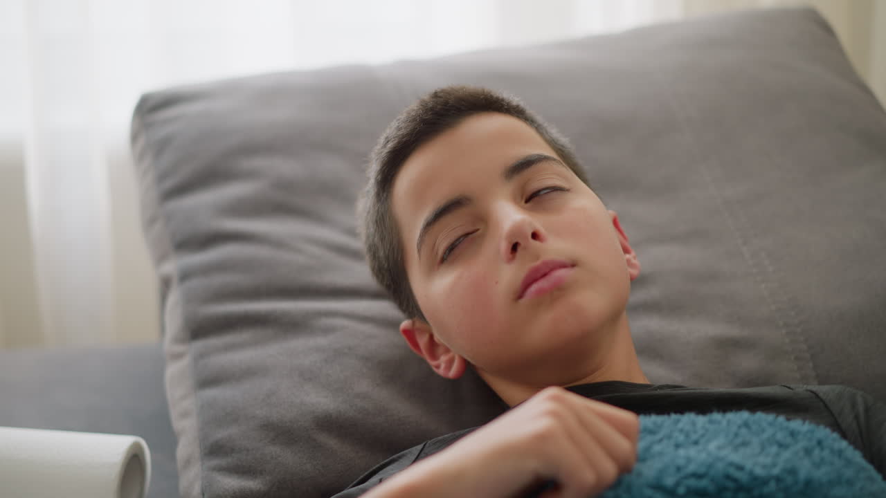 Close-up of sick boy resting on couch covered with blue blanket, coughing, adjusting position while feeling unwell with light streaming through window