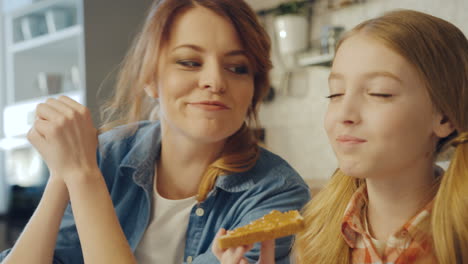 Close-up-of-the-cute-pretty-teen-girl-enjoying-a-sandwich-with-peanut-butter-and-her-mother-looking-at-her,-than-hugging-and-kissing-her.-Portrait-shot.-Indoors