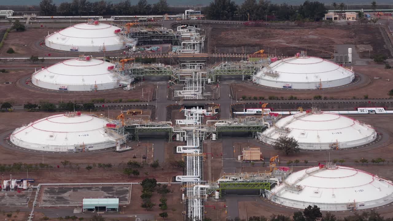 High angle aerial perspective flying over a large liquefied natural gas terminal, showing massive white spherical storage tanks, intricate pipelines, and the surrounding industrial complex