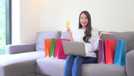 Una-Mujer-Joven-Y-Atractiva-Con-Una-Laptop-De-Rodillas-Sostiene-Una-Tarjeta-De-Crédito-Dorada-En-Una-Mano-Mientras-Señala-Las-Coloridas-Bolsas-De-Compras-Que-La-Rodean-En-El-Sofá