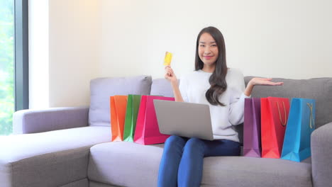 una mujer joven y atractiva con una laptop de rodillas sostiene una tarjeta de crédito dorada en una mano mientras señala las coloridas bolsas de compras que la rodean en el sofá