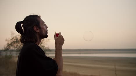 a man with long brunette hair and stubble blows soap bubbles during his vacation outside the city on a summer evening. a mysterious man blows soap bubbles while relaxing near a pond on a deserted shore in the summer evening