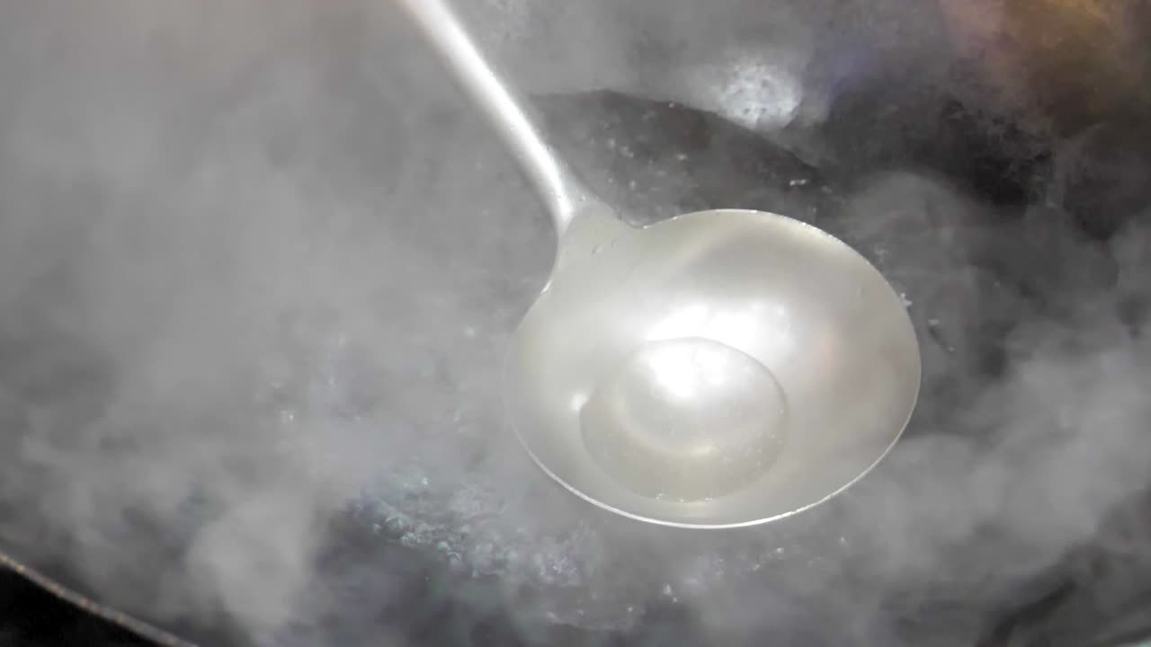 Slow-motion shot of a steaming wok with boiling water, as a metal ladle stirs the bubbling liquid. The intense heat and swirling steam create a dramatic culinary scene.