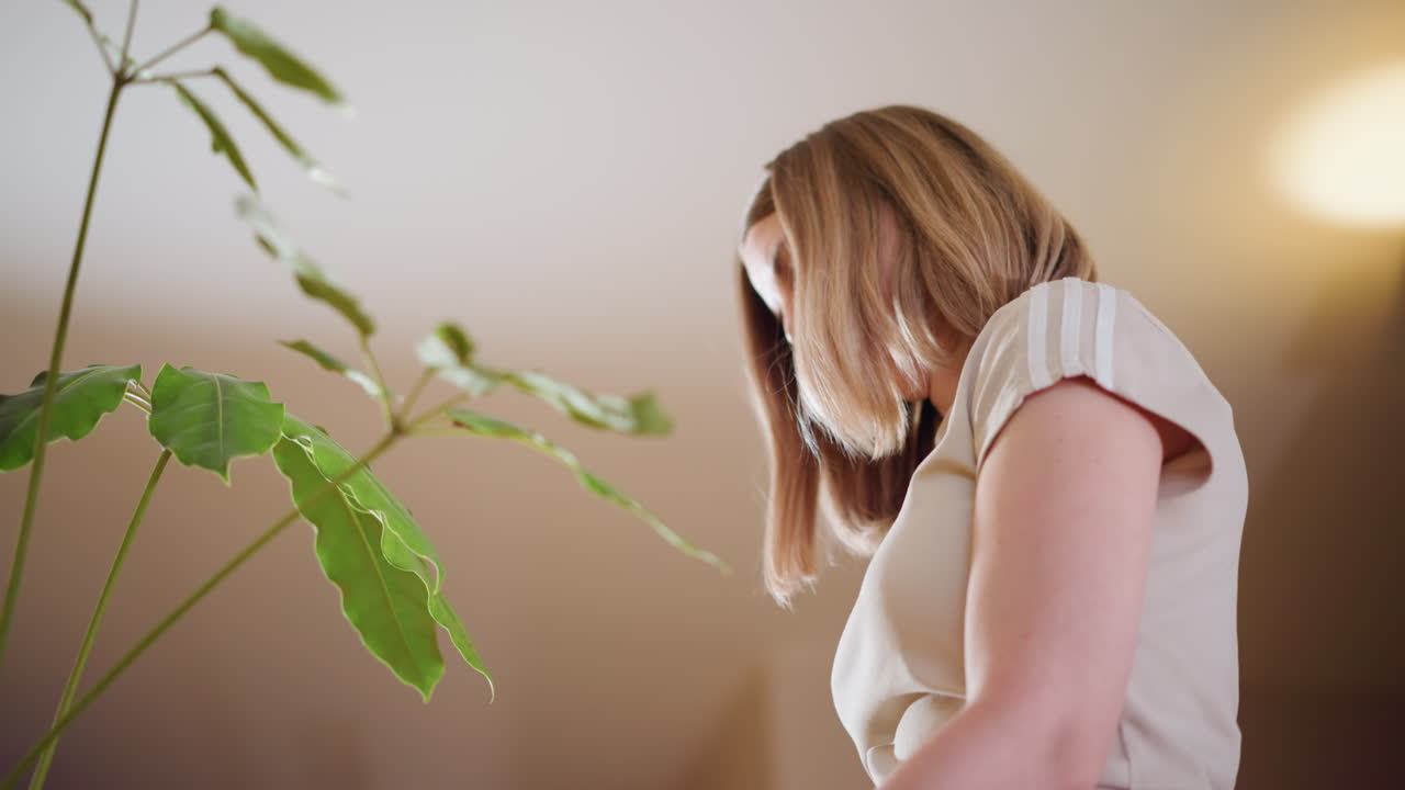 Woman standing near green plant indoors, looking down with soft light highlighting natural atmosphere, representing connection with nature, mindfulness, calm lifestyle, eco friendly values, peaceful home