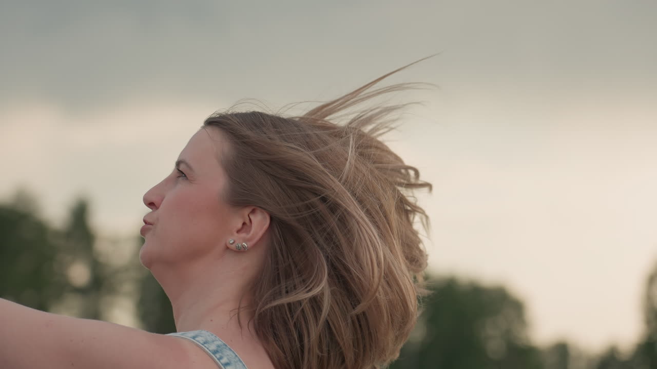 Close up of woman running along asphalt road holding spinning pinwheel fan in hand wind tossing hair against blurred greenery backdrop under summer sky