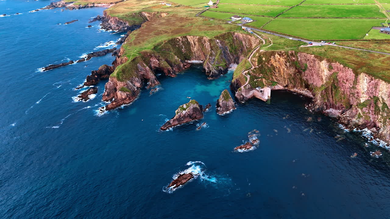 Deep blue waters of the Atlantic Ocean splash by the stunning rocks at the shore. Beautiful scenery of the coastline of Ireland from drone. Multiple cottages are in the valley among the mountains.
