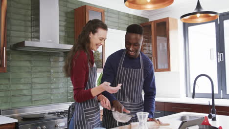 Feliz-Pareja-Diversa-Parada-En-La-Cocina,-Hablando,-Enrollando-Masa-Y-Ensuciándose,-En-Cámara-Lenta