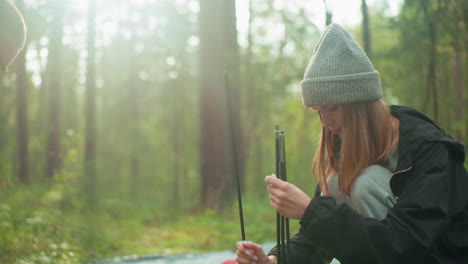 couple preparing tent in tranquil forest as woman aligns multiple tent poles with focus, warm sunlight filters through trees creating gentle glow and peaceful camping atmosphere around them