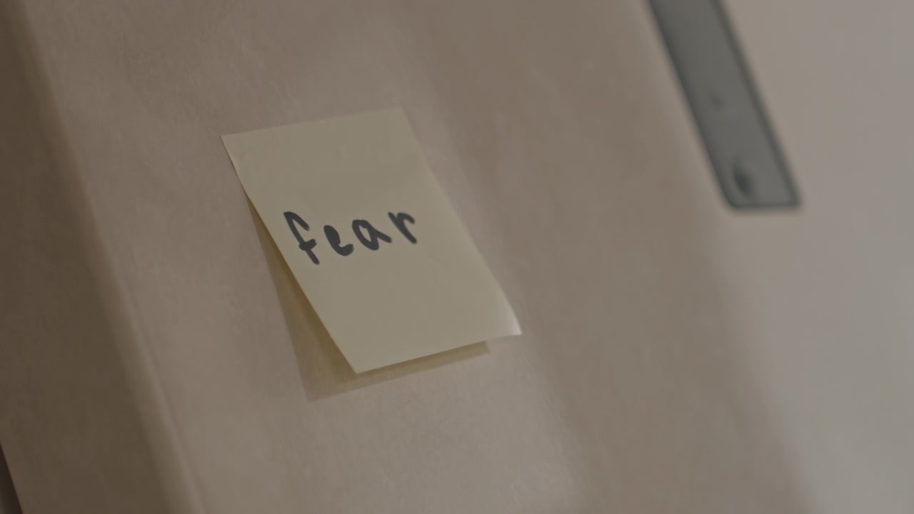 Close up of hands placing sticky note on smooth surface. Sticky note reveals handwritten word fear. Indoor setting with plain background and soft lighting featuring hands