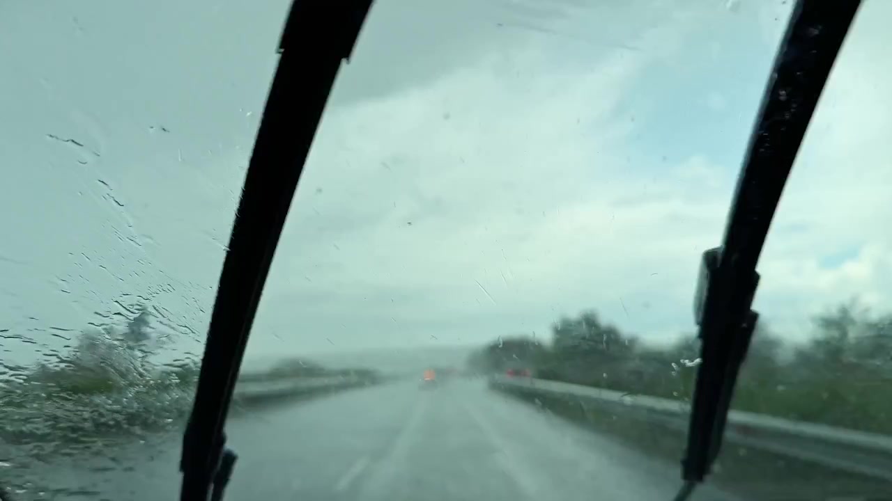 La pioggia cade sull'autostrada mentre un'auto viaggia con i tergicristalli in movimento durante una giornata nuvolosa in una zona suburbana.