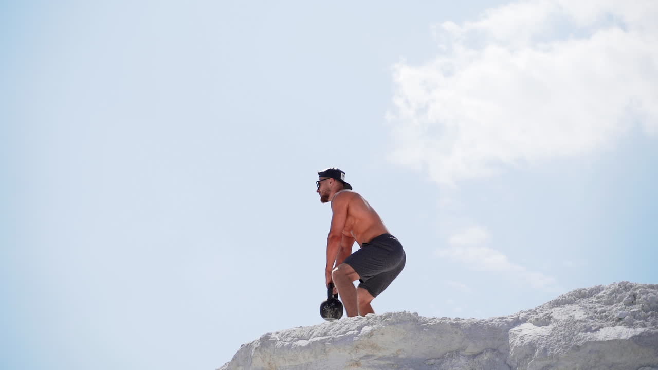 Side view of a strong sportsman training with heavy weight outside. Muscular athlete swings kettlebell on the high hill in a daytime.