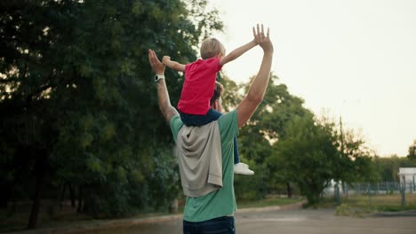 Rear-view-of-a-little-blond-boy-sitting-on-his-dad's-shoulders-in-a-Green-T-shirt,-they-hold-hands-and-raise-them-up.-Happy-little-boy-sitting-on-the-shoulders-of-his-dad-who-is-walking-in-the-park