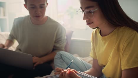 dos adolescentes caucásicos sentados en la cama y aprendiendo de libros y computadoras portátiles.