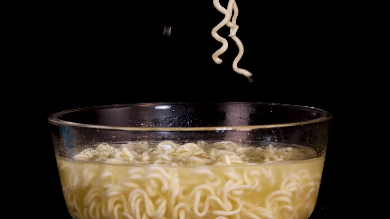 A hand lifts cooked instant noodles from a glass bowl with chopsticks against a black background, highlighting texture, movement, and simple studio lighting
