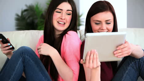 Two-friends-sitting-back-to-back-using-their-smartphone-and-tablet