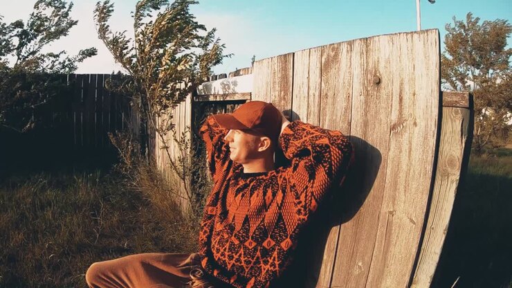 Uomo adulto appoggiato a una recinzione, nel vento, che guarda verso il sole, indossando un maglione nero e rosso.