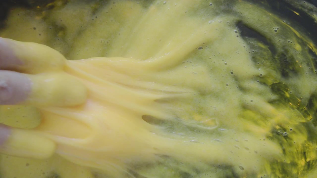 A top-down close-up shot captures a child's hand swirling, pressing, and kneading an elastic mass of bright yellow slime inside a dark tray during a creative home science experiment.
