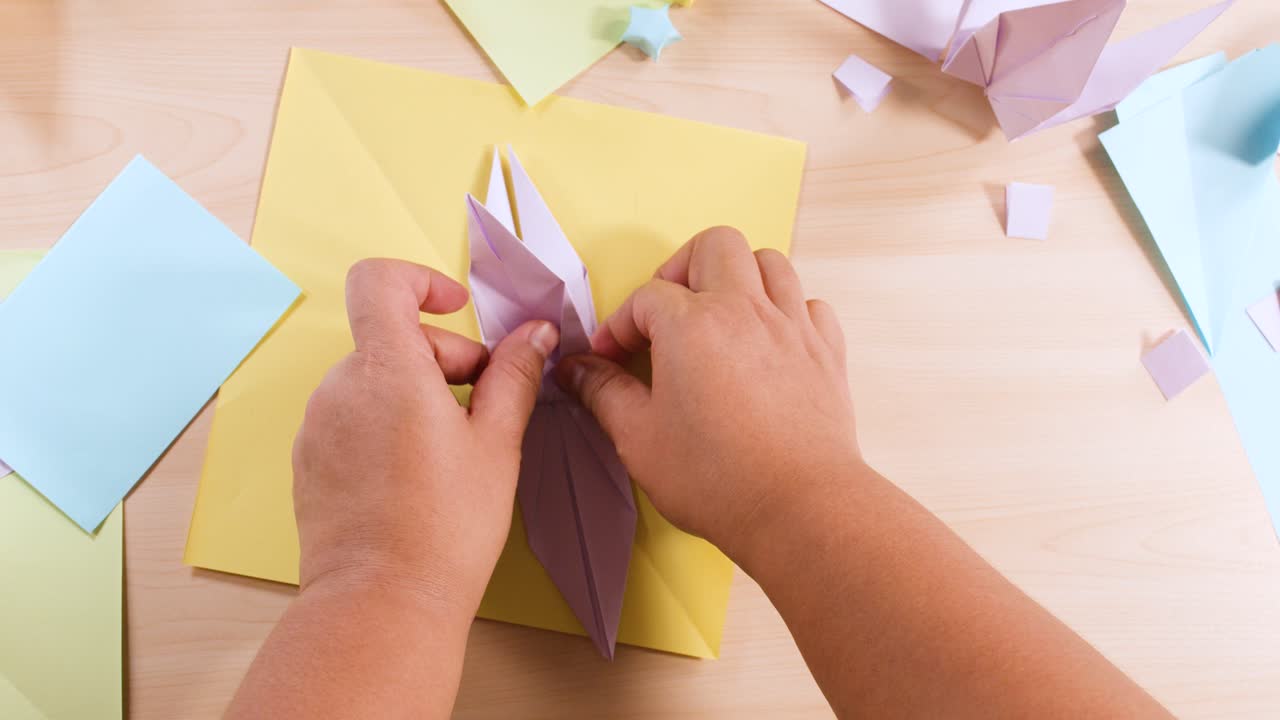 Overhead-billede af hænder, der omhyggeligt folder lilla papir til en origami-fugl på et træbord