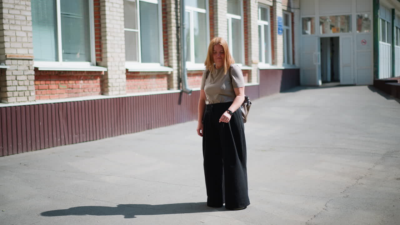 Young lady stands under bright sun near school building, looking around and checking time on wristwatch, wearing casual clothes and fitness bracelet, carrying backpack, waiting thoughtfully outdoors