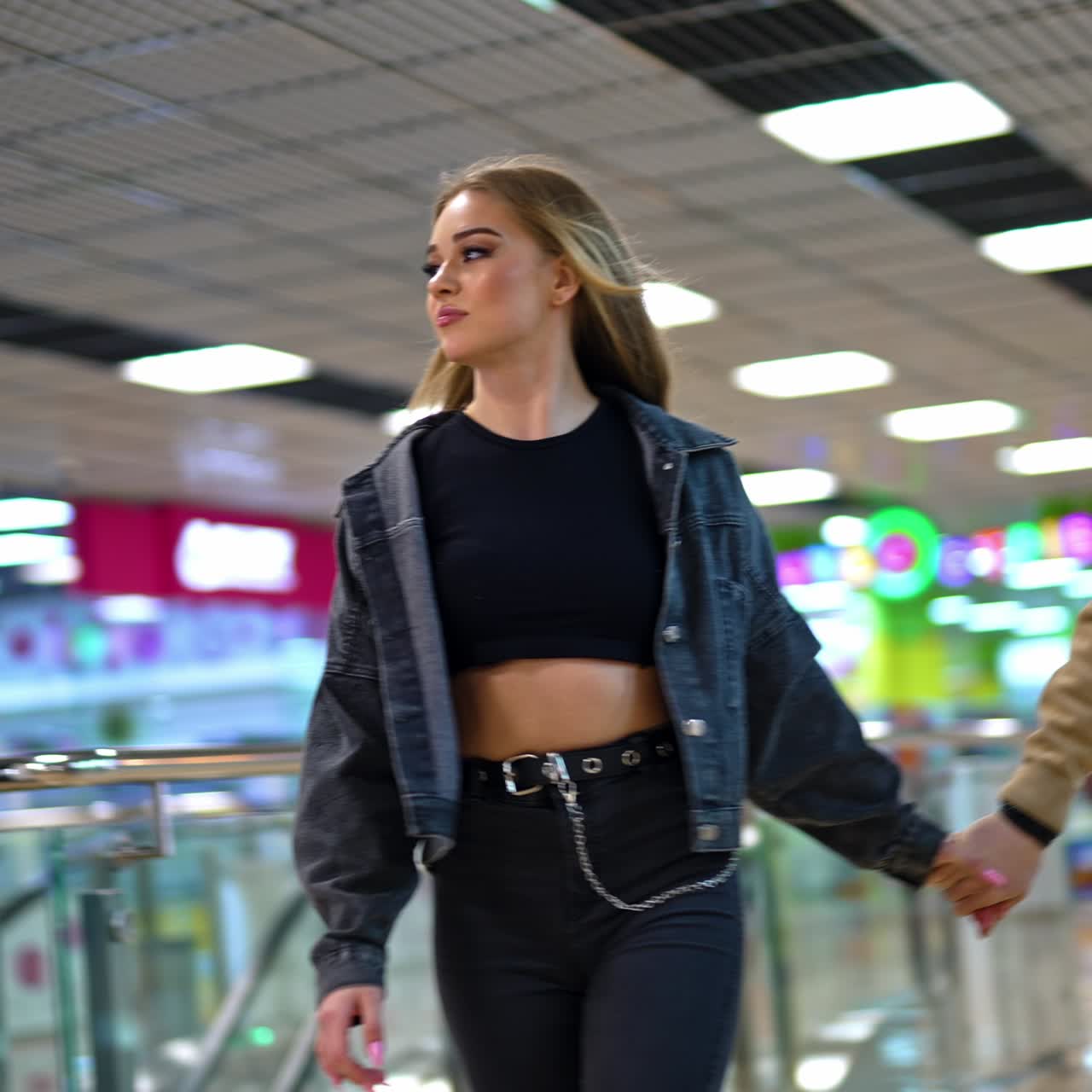 Beautiful young long-haired girl dancing cheerfully. Couple taking by the hands and walk by the shopping mall. Lady sits in a chair of makeup artist