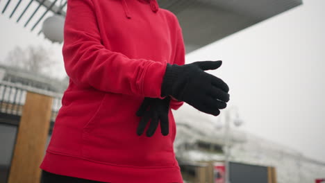 woman putting on black winter gloves for warmth during cold weather, dressed in a red hoodie, background features snowy hill, modern structures, and light pole