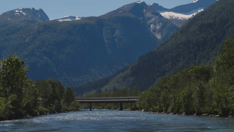 Crystal-blue-river-flowing-towards-a-bridge-in-front-of-tall,-snowy-mountains