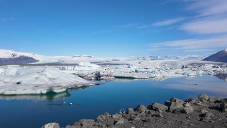 iceland's natural beauty shines in this captivating time-lapse, featuring a serene river, a few swimming ducks, and a picturesque backdrop of snow-covered mountains and clear blue skies