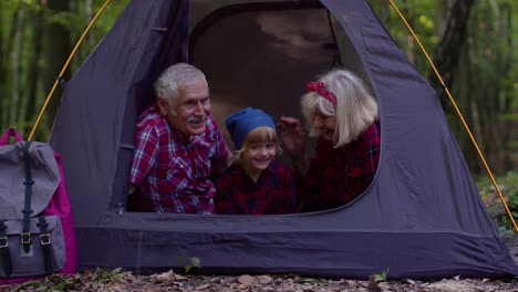 Senior-grandmother-grandfather-with-granddaughter-sitting-in-tourist-tent-over-campfire-in-wood