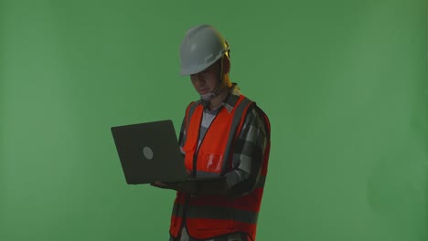 side view of asian male engineer with safety helmet working on a laptop while standing in the green screen background studio, during sunset or sunrise time