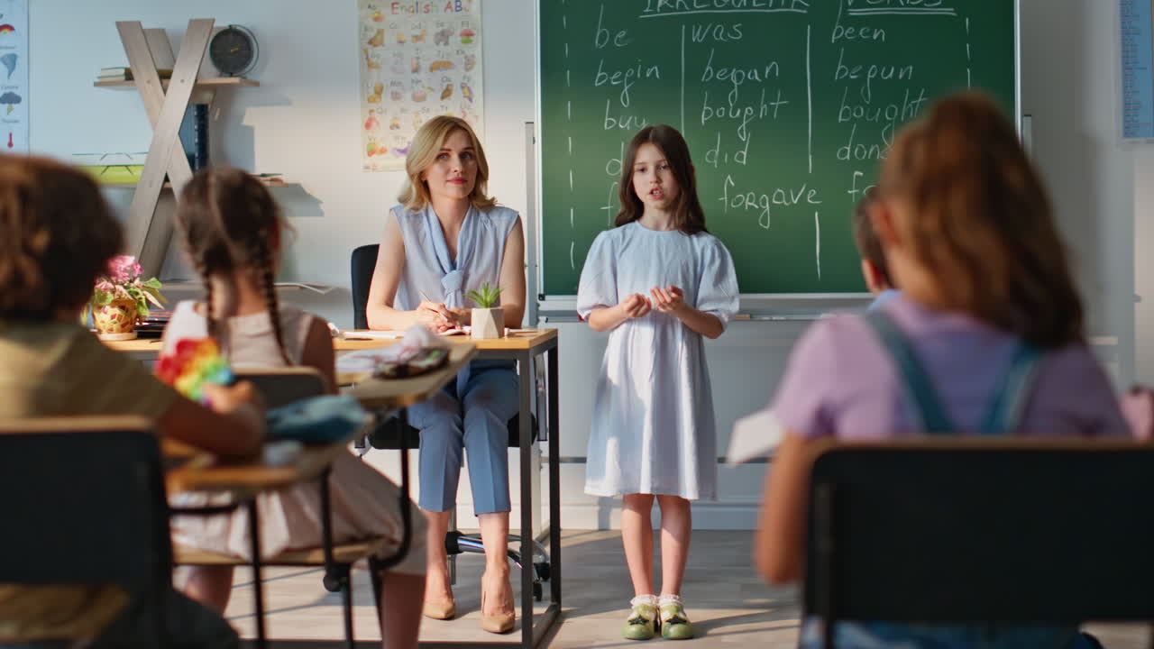 Teacher interacting little girl standing at classroom blackboard. Cute pupil