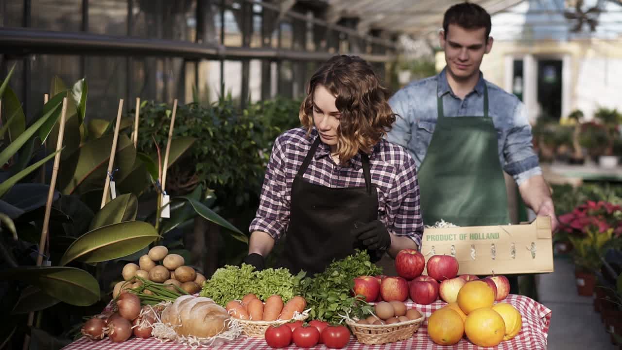Slow motion footage of a handsome, tall man in green apron carrying the box with fresh unrecognizable vegetables and fruits to the woman selling organic products. She counts harvest and making notes. Indoors