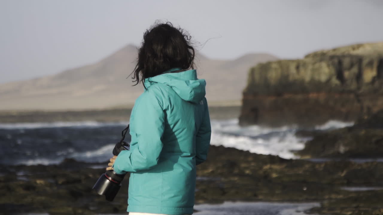 Donna turista in piedi su una scogliera rocciosa sull'oceano con la sua moderna fotocamera a Fuerteventura, Isole Canarie, Spagna, meta turistica, paesaggio marino naturale tropicale