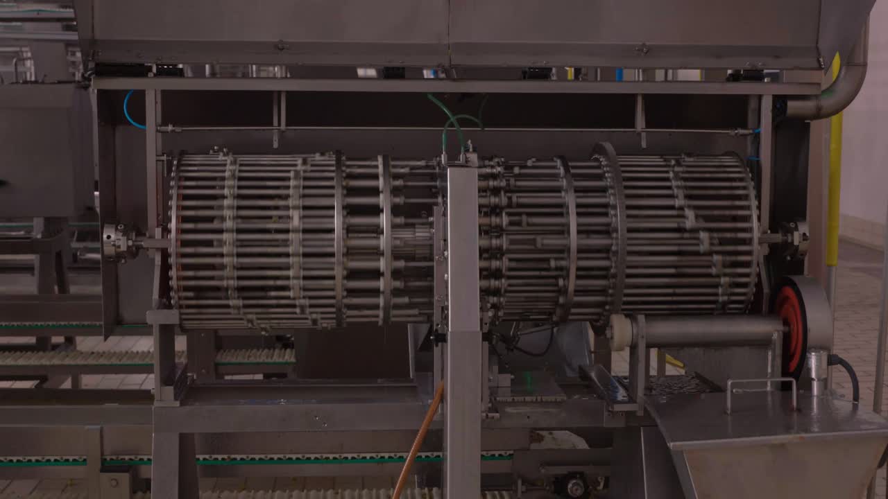 A slow-motion zoom-in shot focuses on a metallic food processing machine, revealing its intricate cylindrical components and green tubing in a factory environment.