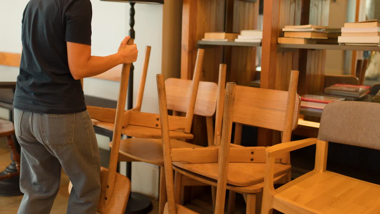 A person stacks wooden chairs against a bookshelf in a warm, indoor restaurant setting