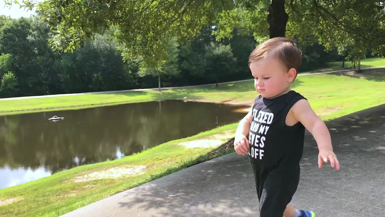 Toddler walking down a path beside a small pond with woods in background.