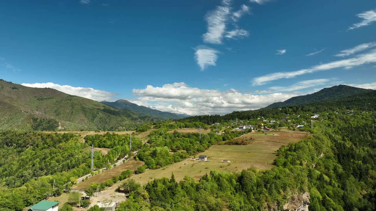 A wide aerial shot of a hidden valley in the Georgian Caucasus, surrounded by endless green slopes and a few scattered houses under a bright summer sky