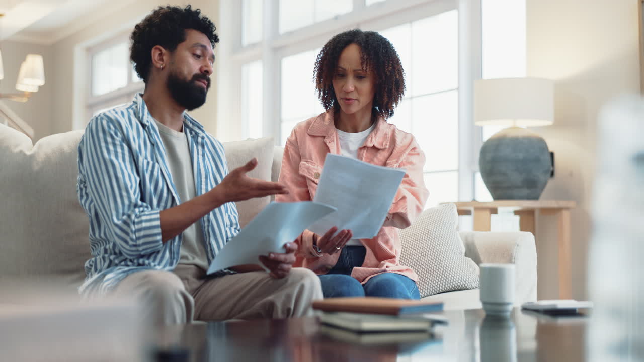 A couple sitting on a sofa in a living room, looking at papers and talking.