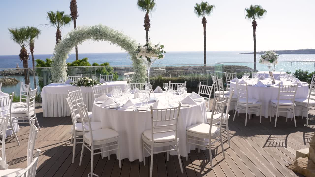 Beautiful outdoor wedding setup with round tables dressed in white linens. A floral arch frames the stunning sea view, creating a perfect atmosphere for a celebration by the shore