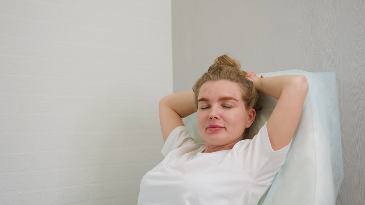 Blonde woman sitting on salon chair with hands supporting head, eyes closed, appearing relaxed against soft-lit gray and white walls. Scene suggests calm, comfort, and personal downtime