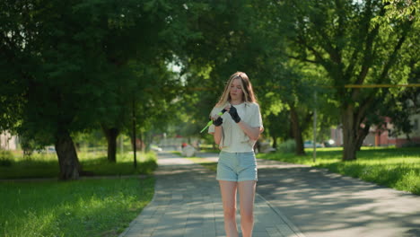 young woman strolls through sunlit park, observing green air pump in her hands with concentration, sunlight reflects off her, while people move in background among lush trees and distant buildings