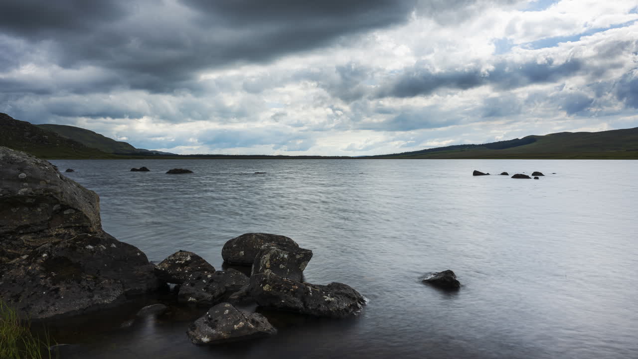 lapso de tiempo del lago con hierba y grandes rocas en primer plano en un oscuro día de verano nublado en el paisaje rural de irlanda