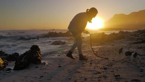 mujer joven juega con el perro jack russell en la playa durante la hermosa puesta de sol, pequeñas olas rompiendo en la playa rocosa