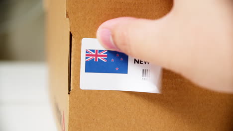 manos aplicando etiqueta de bandera hecha en nueva zelanda en una caja de cartón de envío con productos