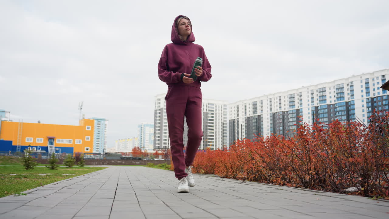Young female athlete in maroon hoodie strolls along paved city park walkway clutching water bottle, enjoying overcast morning, modern skyline and fiery autumn shrubs framing urban fitness routine