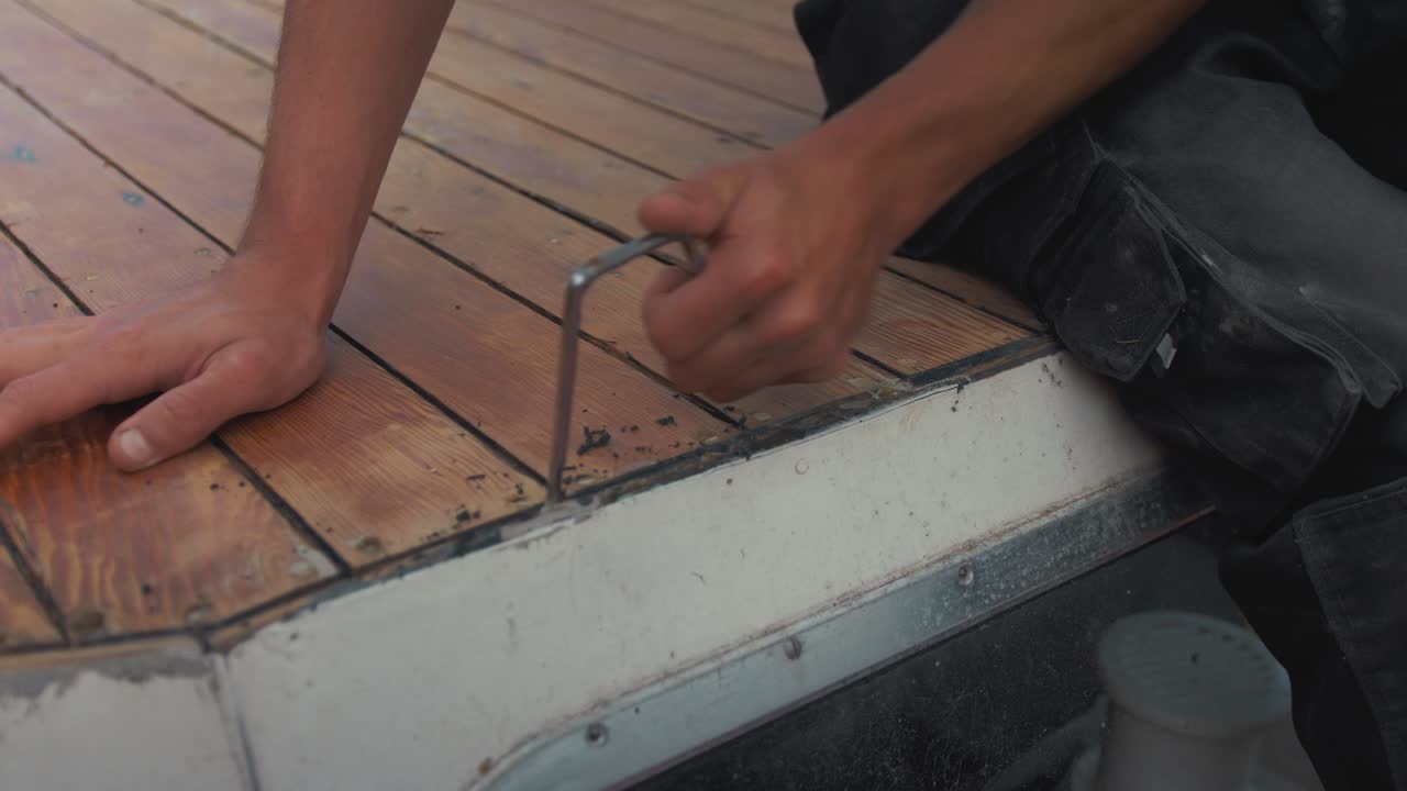Raschiatura di vecchio sigillante in mastice tra le assi del ponte di una barca in legno.