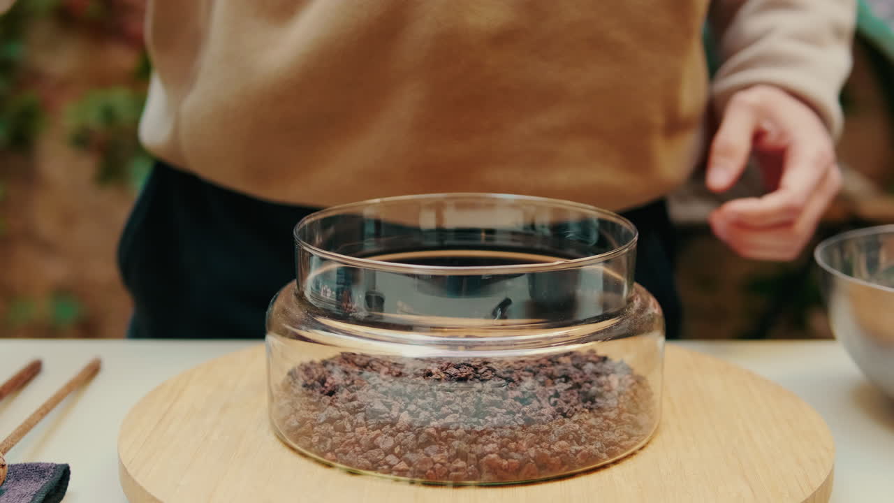 Close-up of Hand Pouring Small Brown Granules into Glass Container