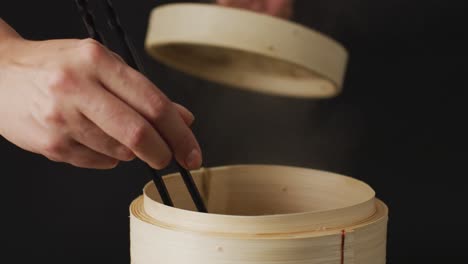 Hand-holding-chopsticks-with-gyoza-dumpling-in-bamboo-steamer-on-black-background