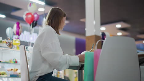 close-up of young woman sitting at mall table reading book, with colorful shopping bags on side, bright background with toy store, balloons, vibrant mall atmosphere